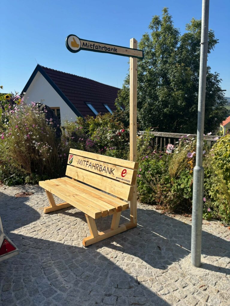 Mitfahrbank mit Daumen hoch-Symbol und Gemeindewappen in einer blumenreichen Umgebung